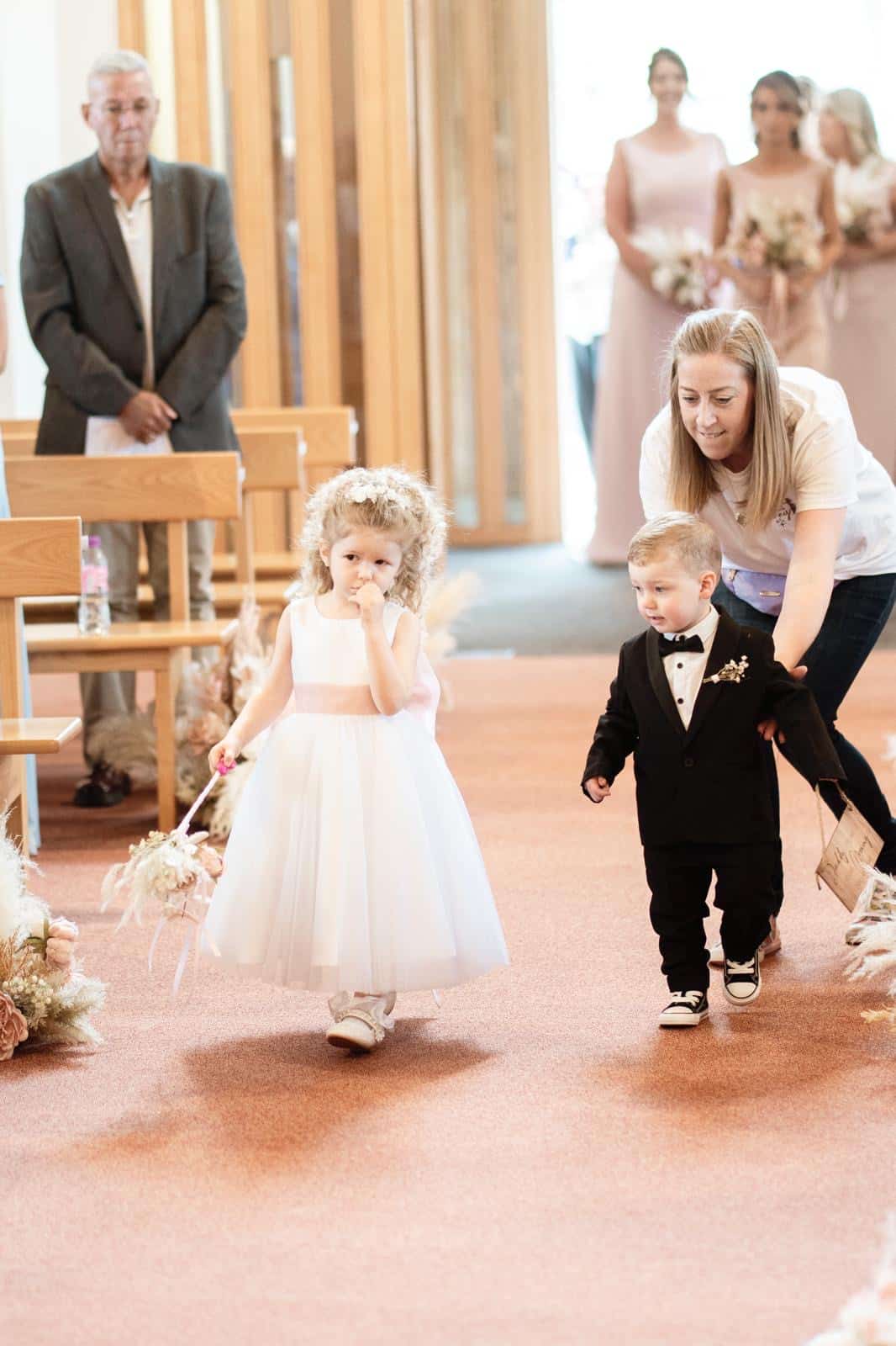 Professional wedding nanny gently guiding a flower girl and page boy down the aisle during a Northern Ireland wedding ceremony, supporting them as they carry out their special roles.