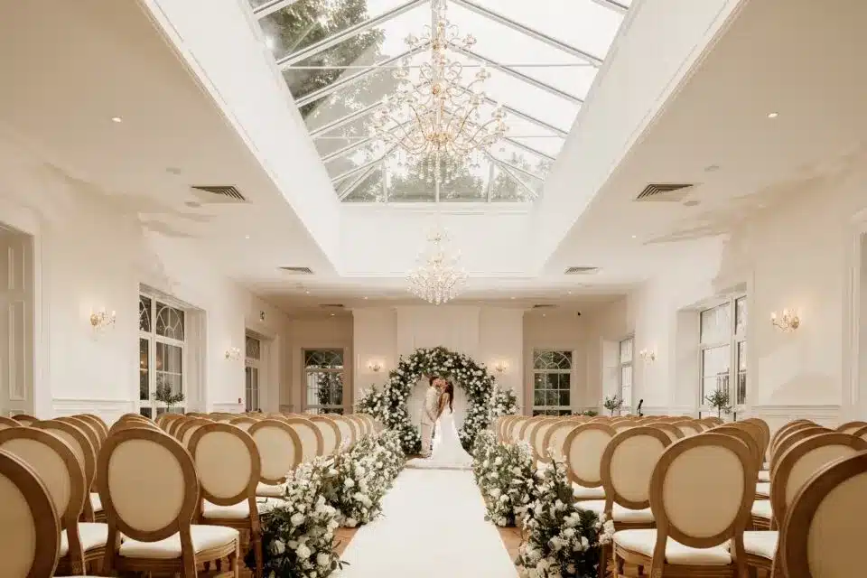 Bright indoor wedding ceremony aisle with floral decorations and seating.