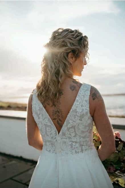 Bride with floral tattoos in lace wedding dress stands outdoors by water.