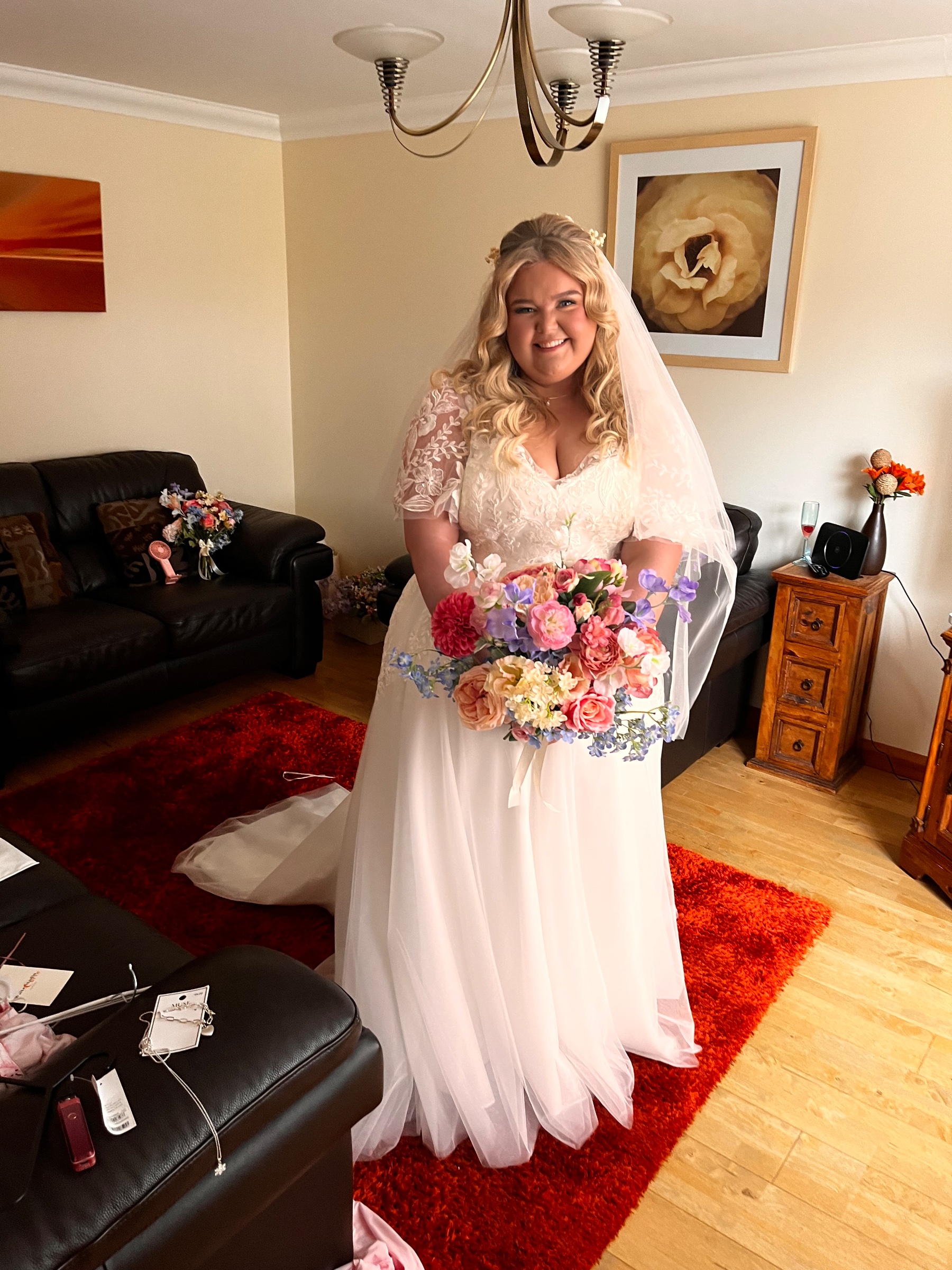 Smiling bride in an elegant wedding dress holding a bouquet of red and white flowers at a reception.