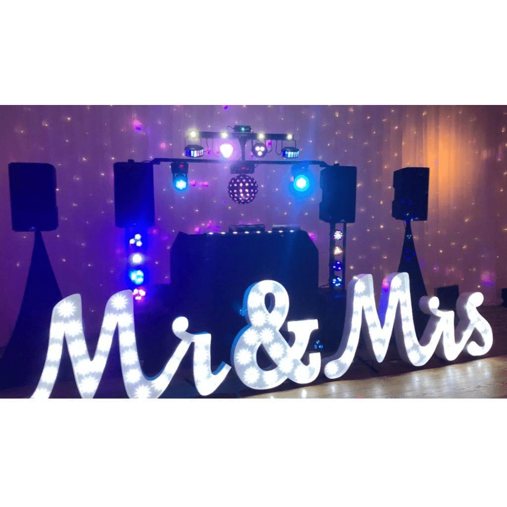 Illuminated "Mr & Mrs" sign with DJ setup and colorful lights at an event.