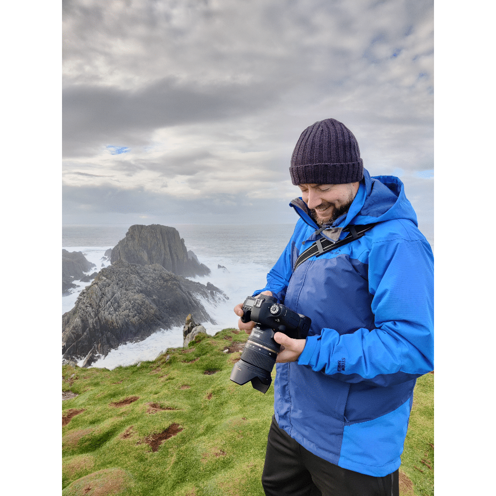 Man in a blue jacket and beanie holding a camera near a rocky coastal landscape.