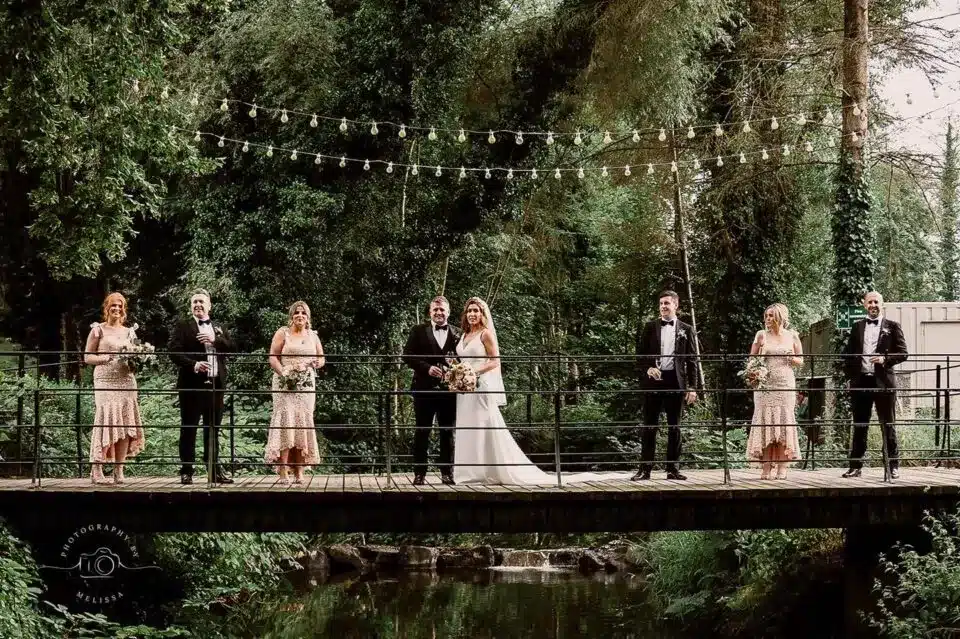 Outdoor wedding ceremony with couple and guests under string lights in wooded setting.