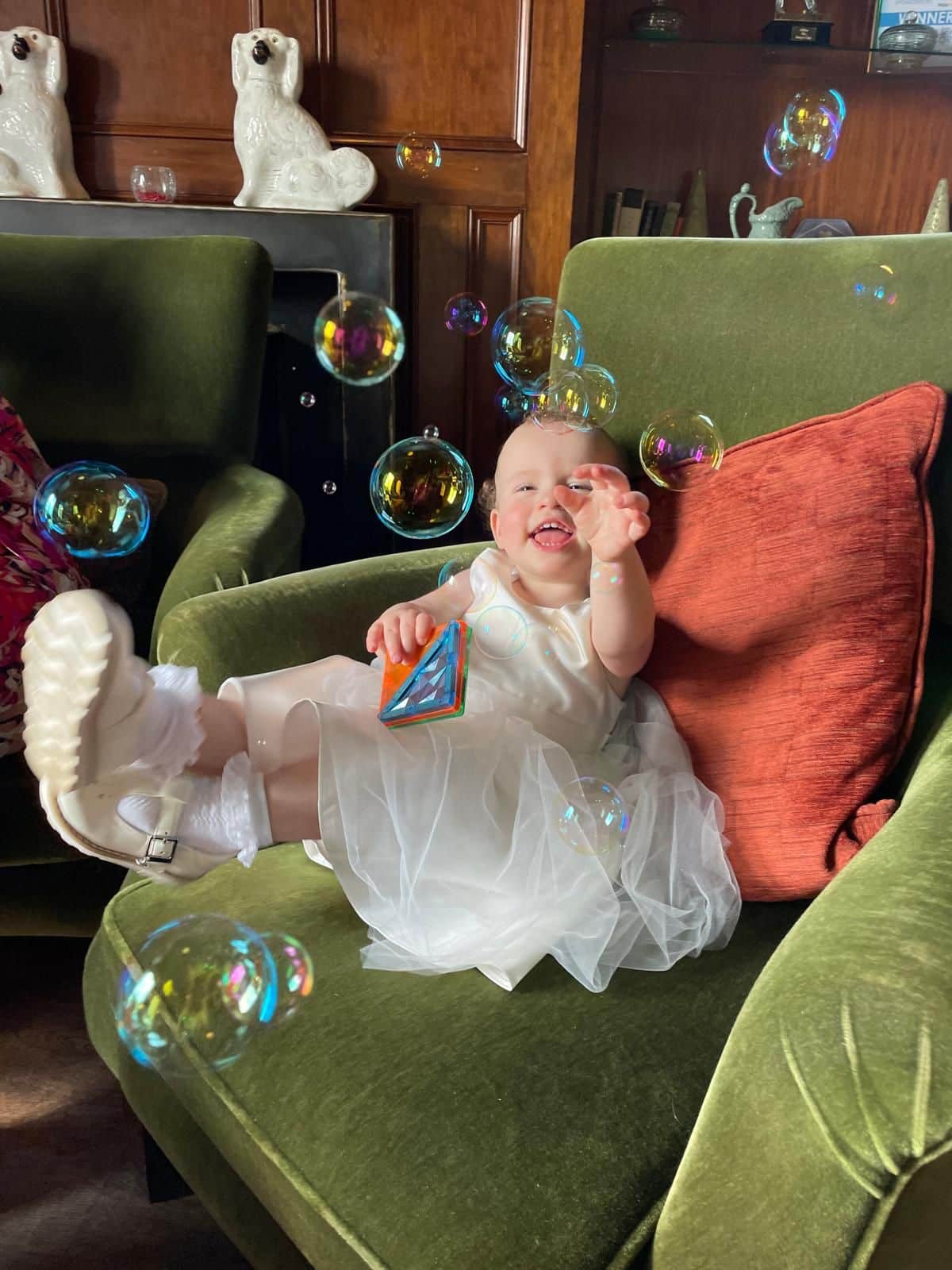 Baby girl in a white dress sitting on a green armchair at a wedding venue, laughing and reaching for bubbles during drinks reception in Northern Ireland.