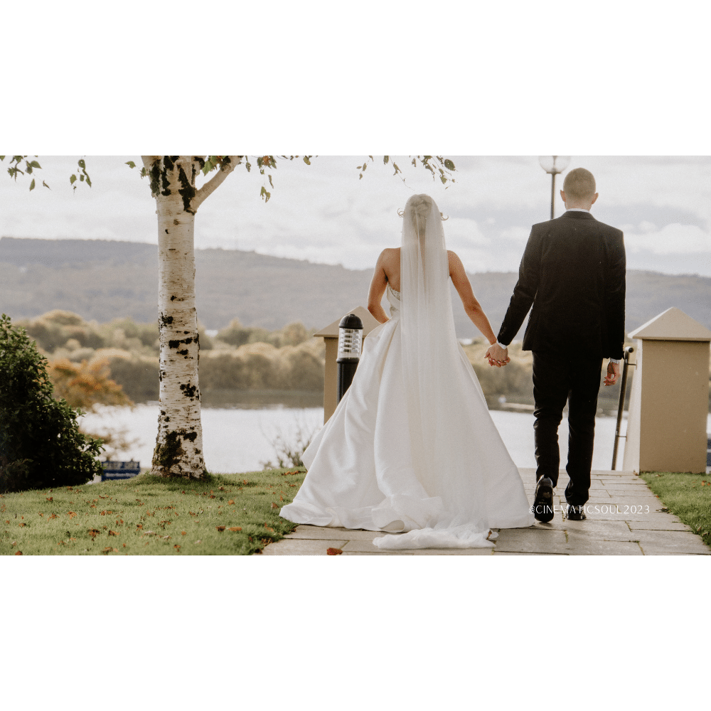 Bride and groom holding hands, walking towards a lake with lush green surroundings.