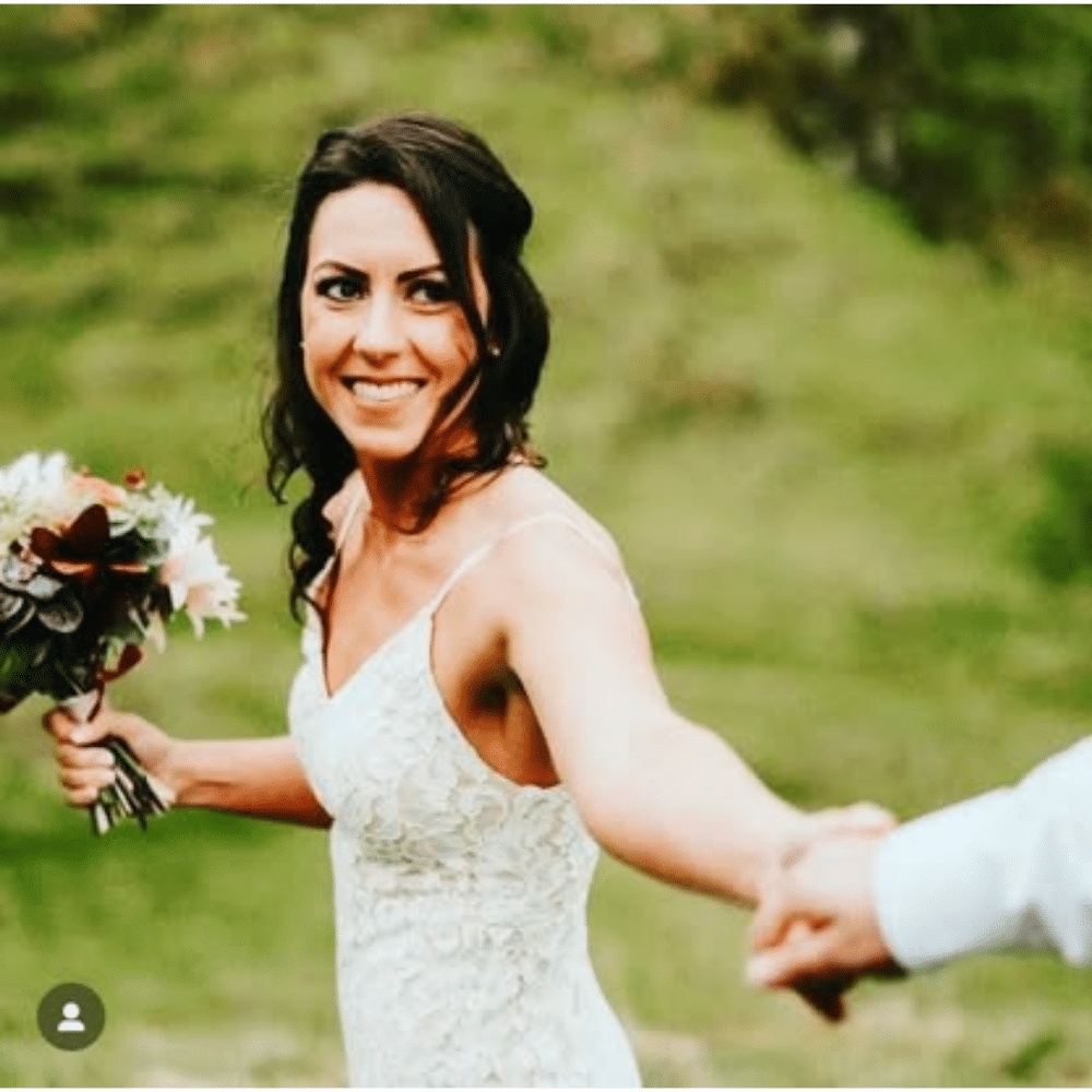 Bride in white dress smiling and holding hands with someone, standing against a green outdoor background.