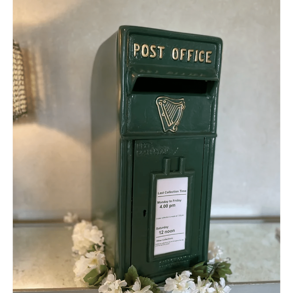 Green vintage post office box with a harp emblem and collection times, surrounded by white flowers.