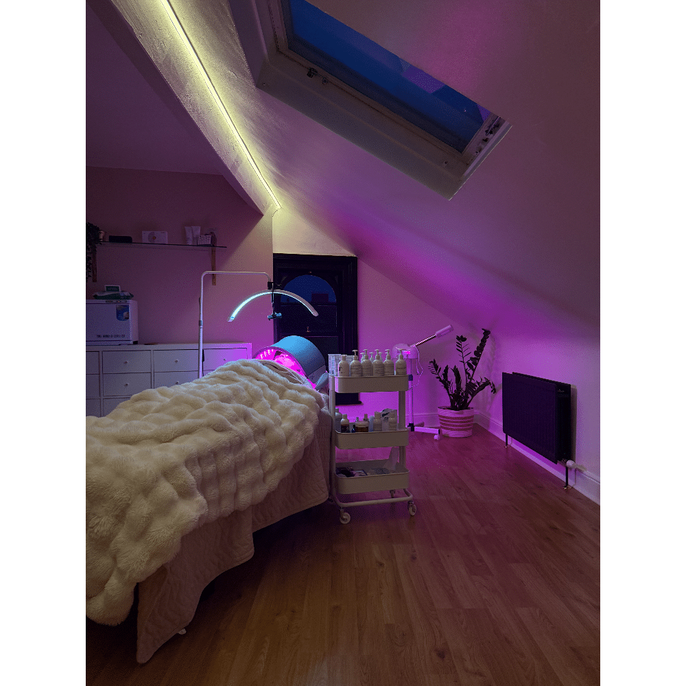 Cozy spa room with treatment table, fur blanket, LED lights, and skincare products under a skylight.