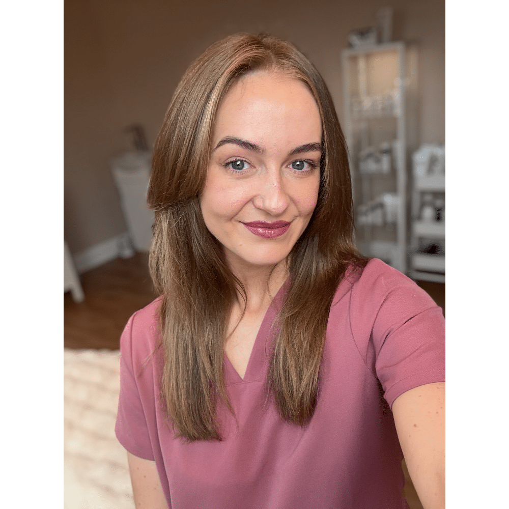 Smiling person with long brown hair in a pink top indoors.