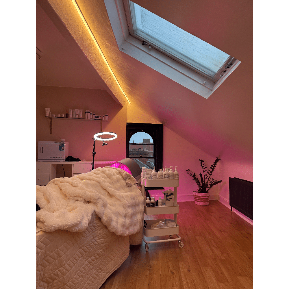 Cozy attic spa room with skylight, treatment bed, ring light, and skincare products on a cart, illuminated by ambient lighting.
