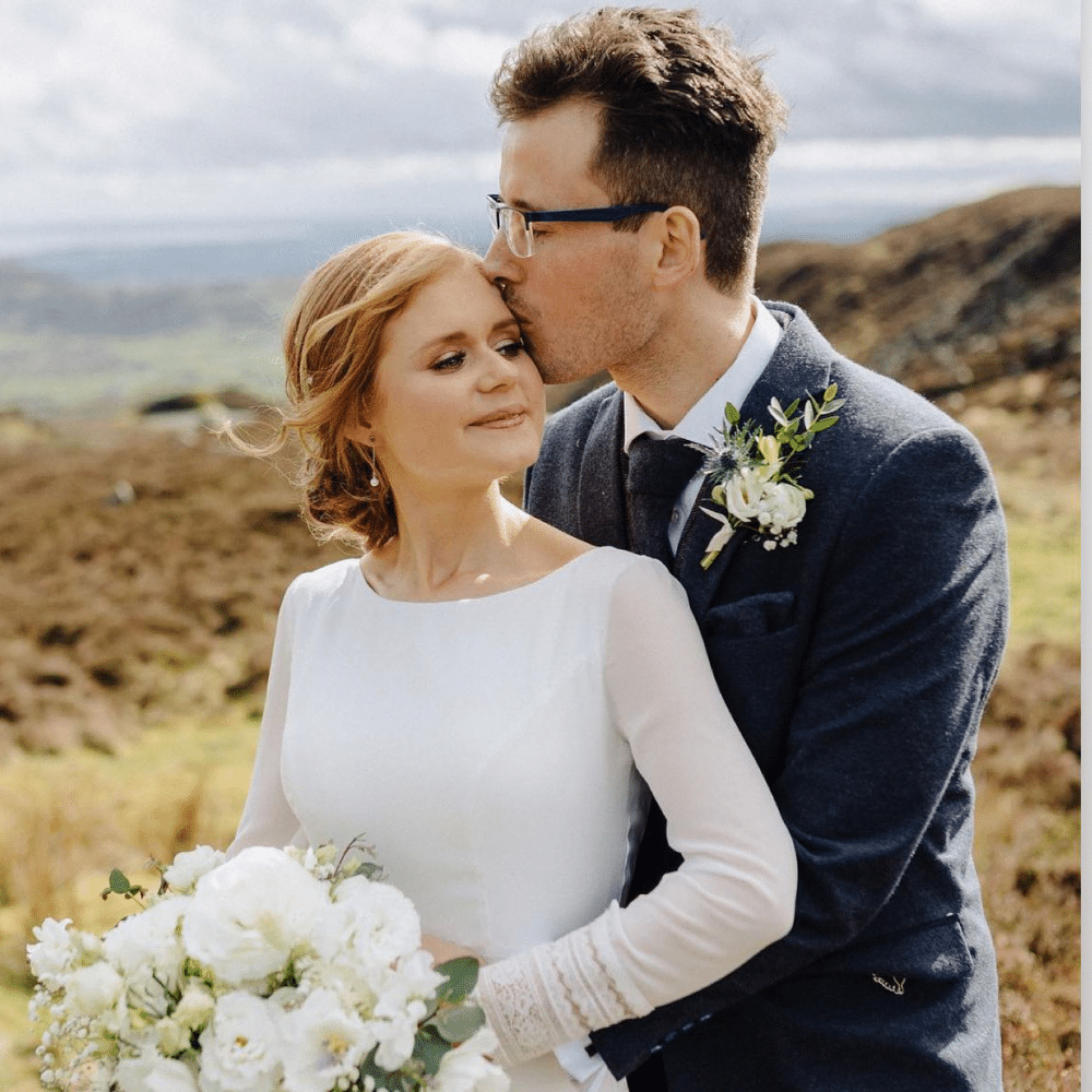 Bride and groom embrace in scenic outdoor wedding photo, with groom kissing bride's forehead.