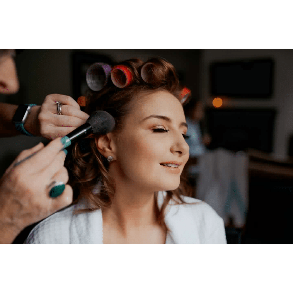 Woman getting makeup applied, wearing hair rollers, and smiling.
