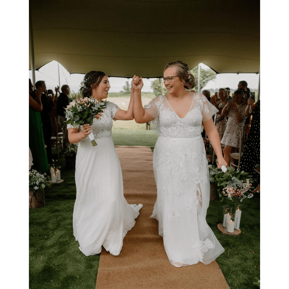 Two brides walking down the aisle, smiling and holding hands, celebrating their wedding in an outdoor ceremony.