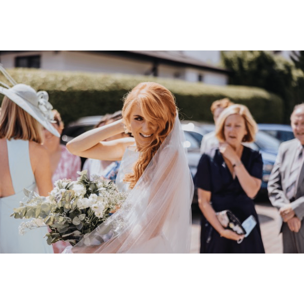 Bride smiling and holding bouquet outdoors with guests nearby.