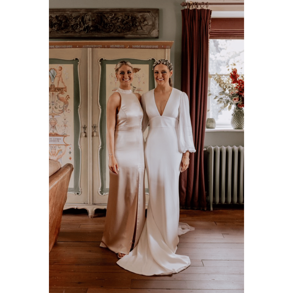 Two women in elegant dresses posing indoors, with floral decor in the background.