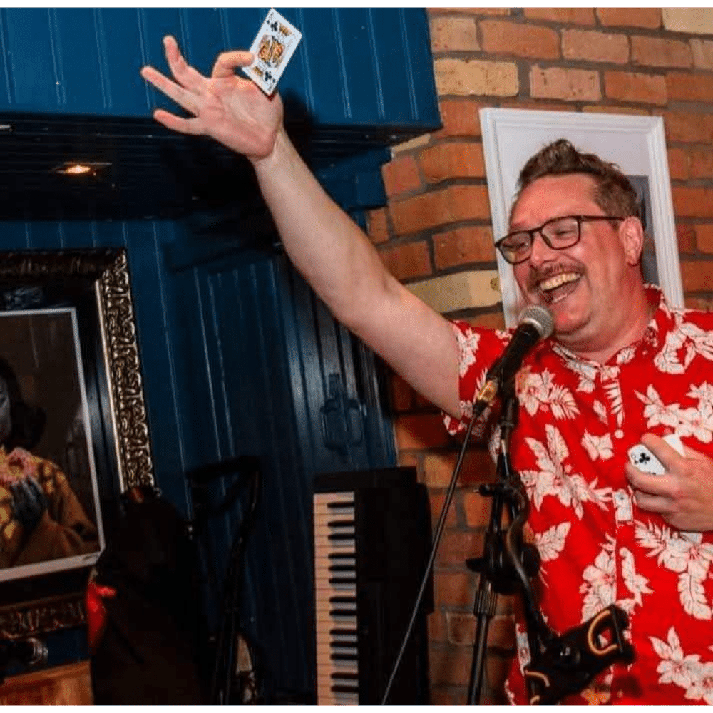 Magician performing card trick on stage in floral shirt, holding card and microphone.