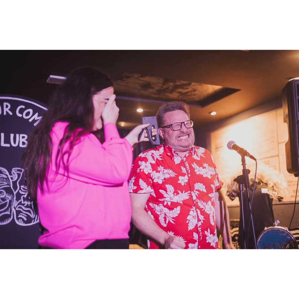 Two people at a comedy club: one holds a phone, the other wears a floral shirt, both appear amused.