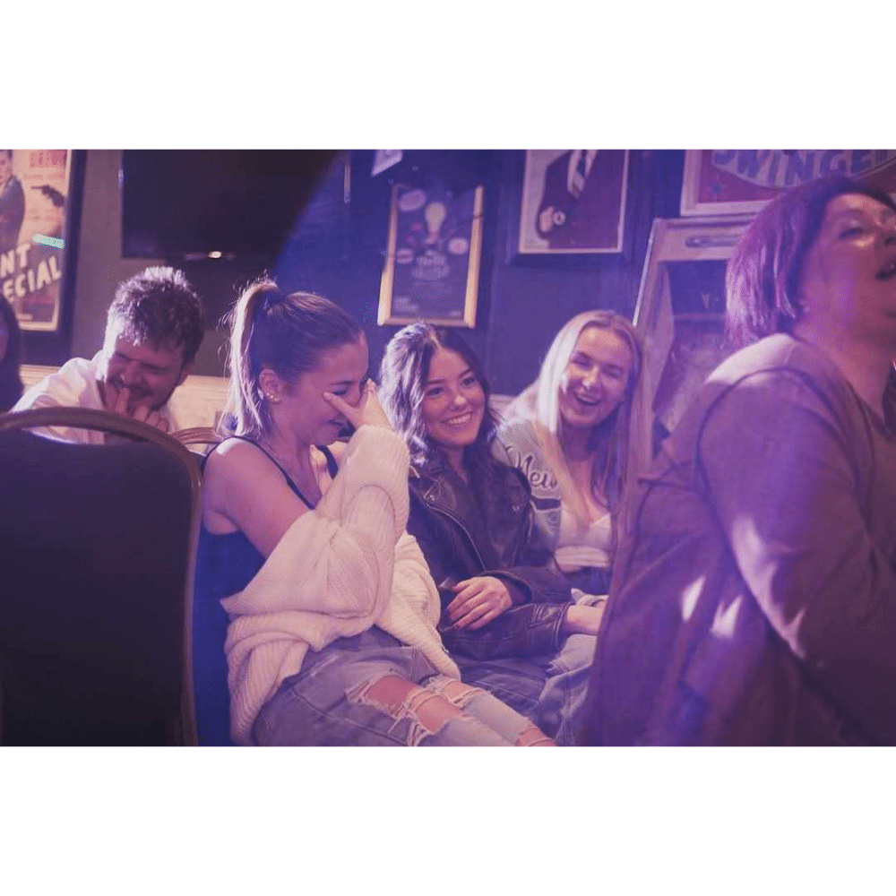 People laughing and enjoying a comedy show in a dimly lit venue with posters on the wall.