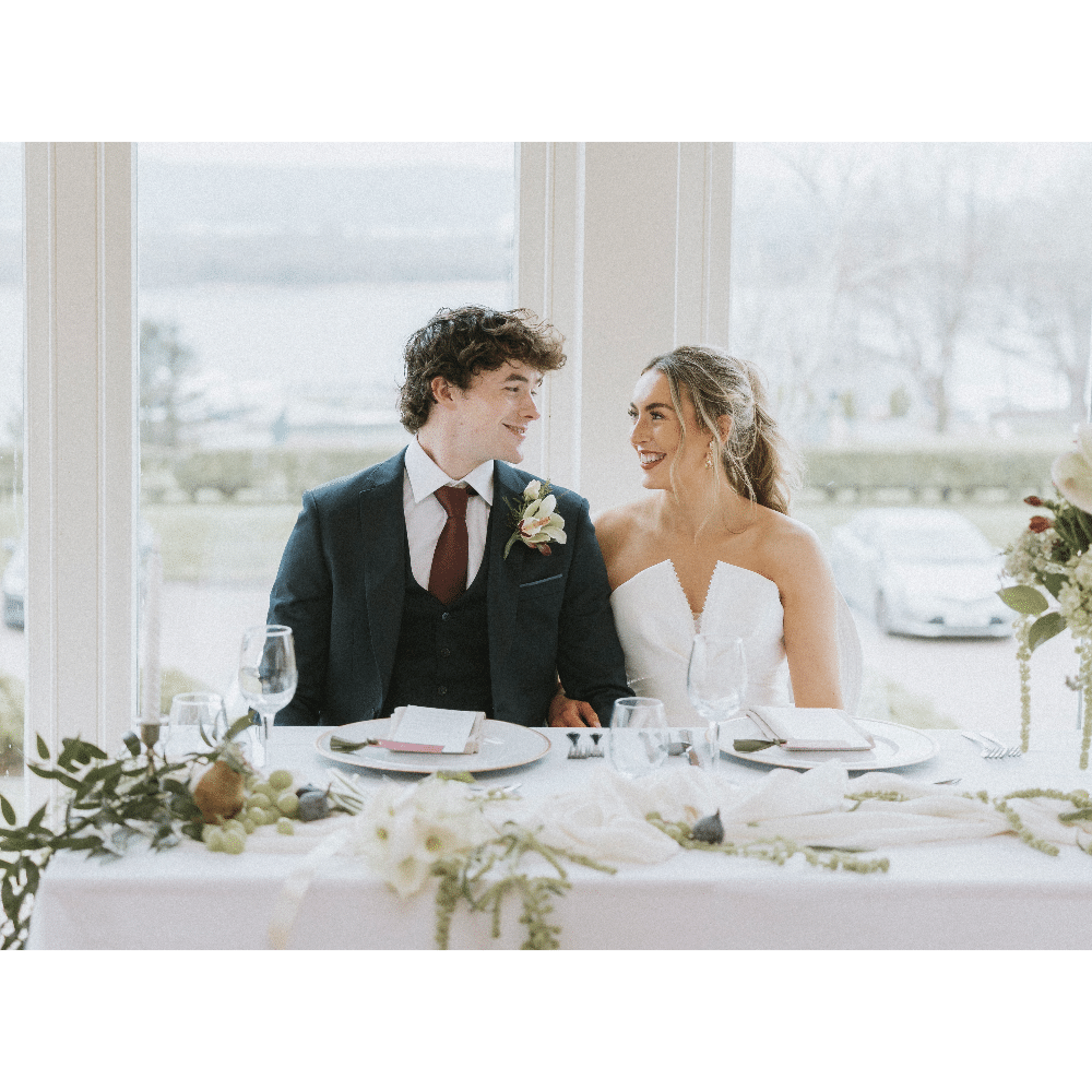 Bride and groom smiling at each other at a decorated wedding table indoors.