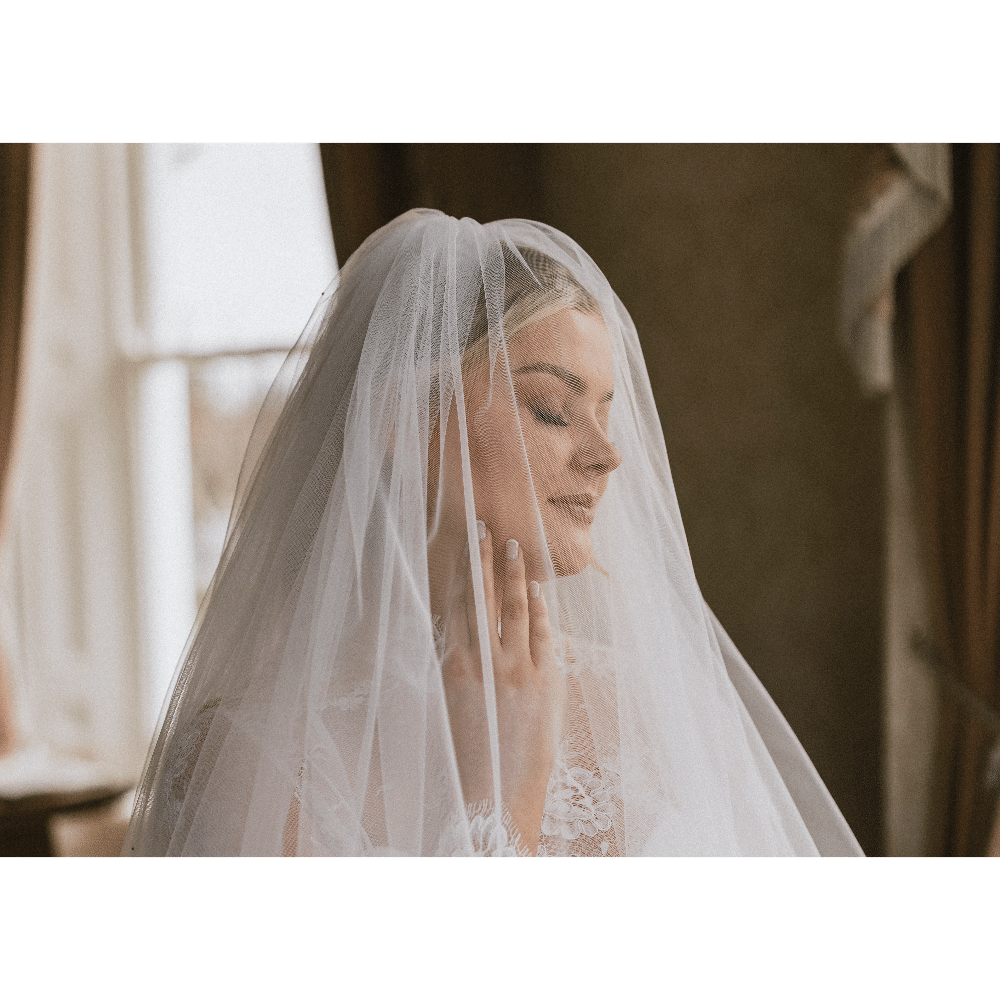 Bride in lace dress and veil, eyes closed, standing near a window.