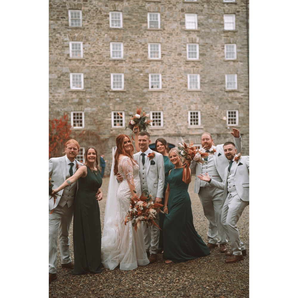 Wedding party posing joyfully against a stone building, bride and groom in center, bridesmaids in green and groomsmen in gray suits.