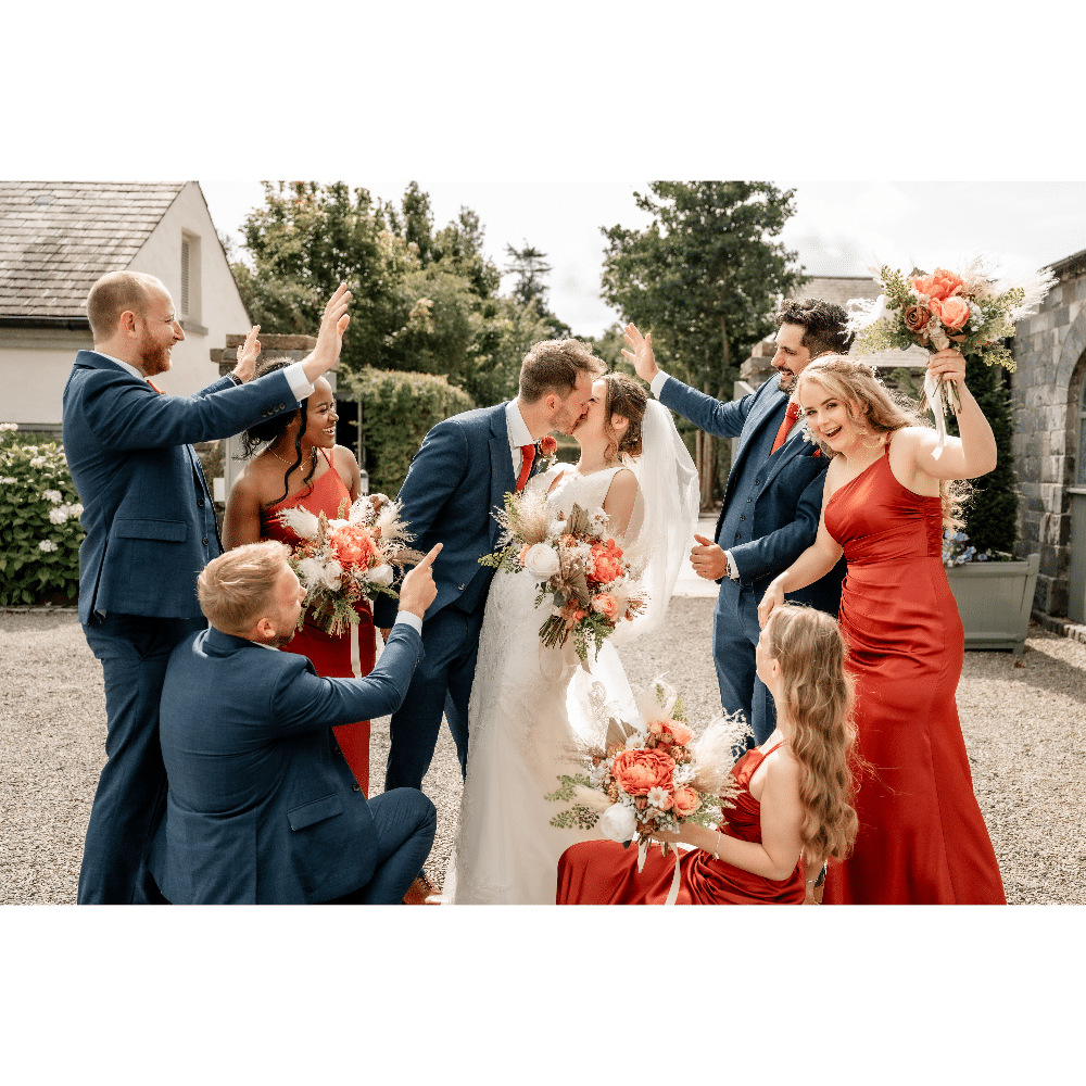 Bride and groom kissing surrounded by wedding party in red dresses and blue suits celebrating outdoors.