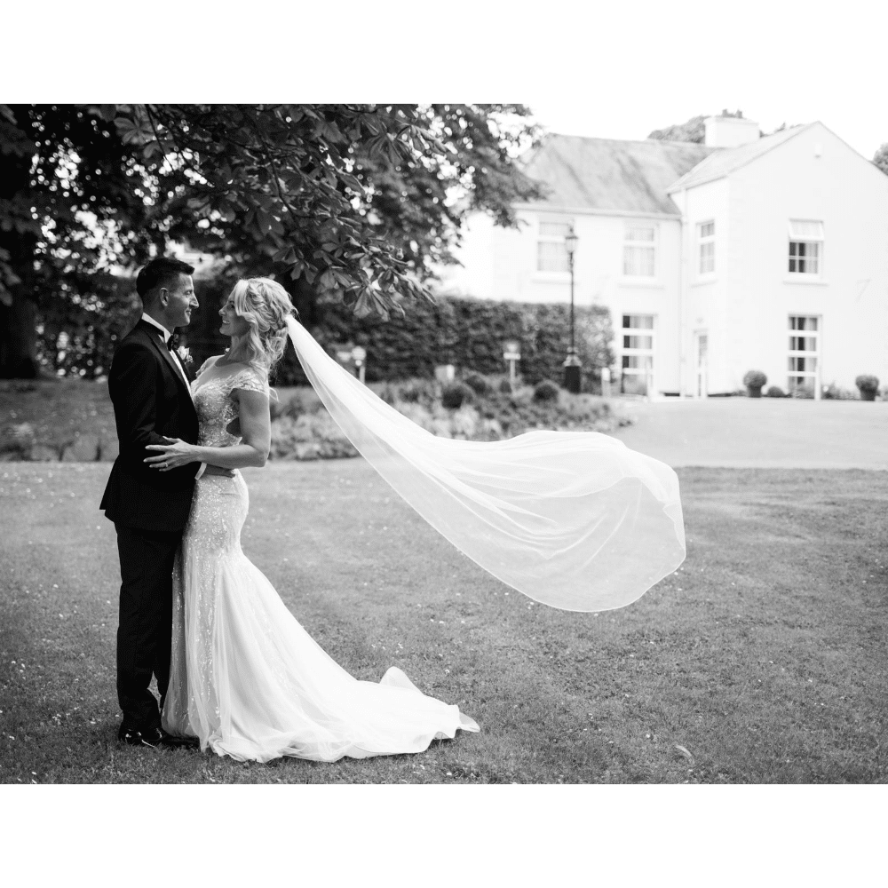 Bride and groom embrace outside, her veil flowing in the wind, with a white house in the background.