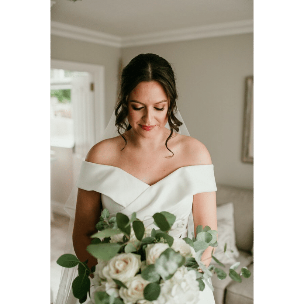Bride in off-shoulder white dress holding a bouquet of white roses and greenery indoors.