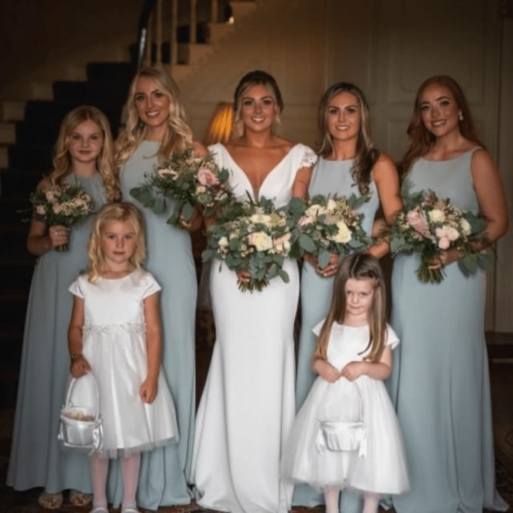 Bridal party group photo with bridesmaids in blue dresses and flower girls in white, holding bouquets indoors.