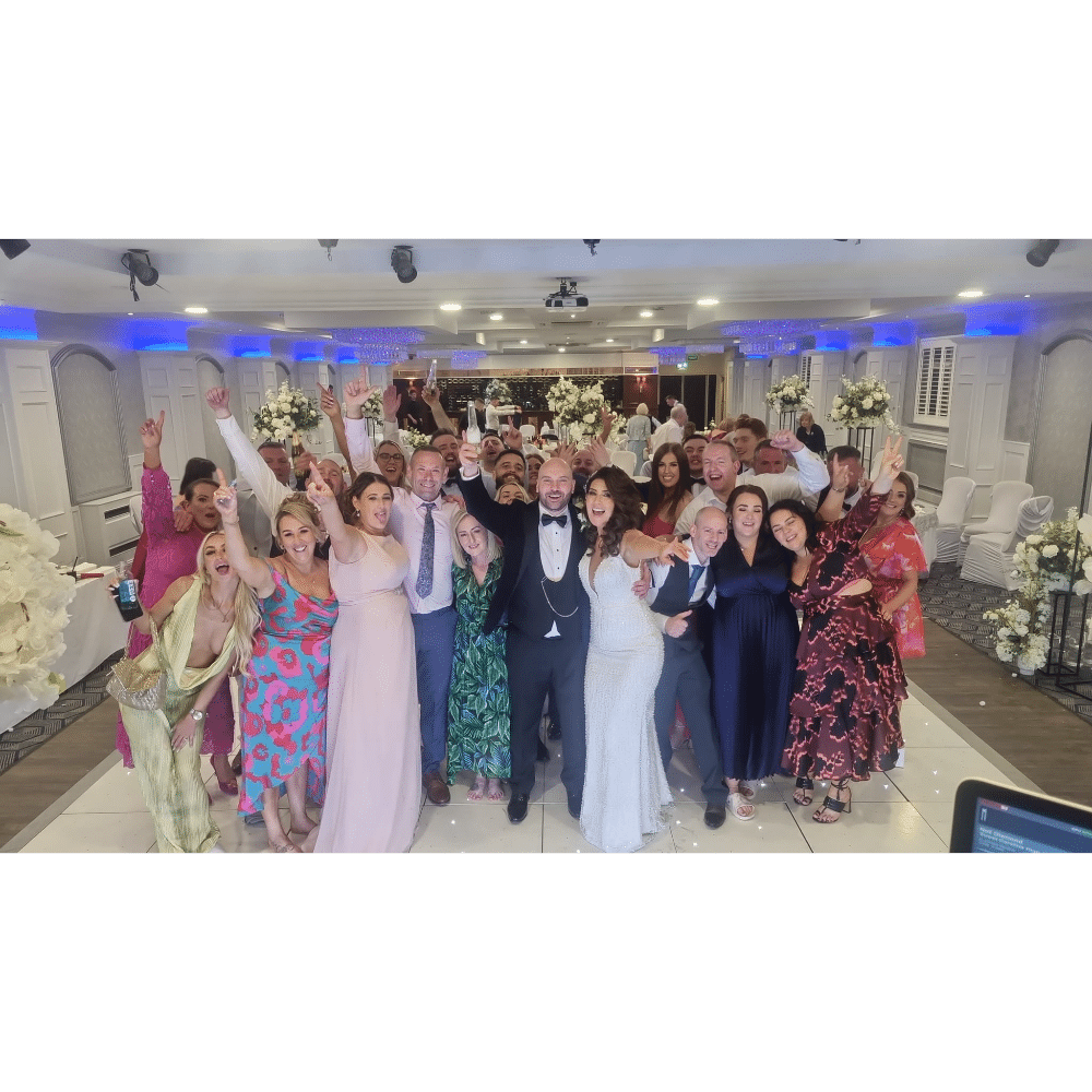A large group of people celebrating at a wedding, including the bride and groom in the center, in a decorated banquet hall.