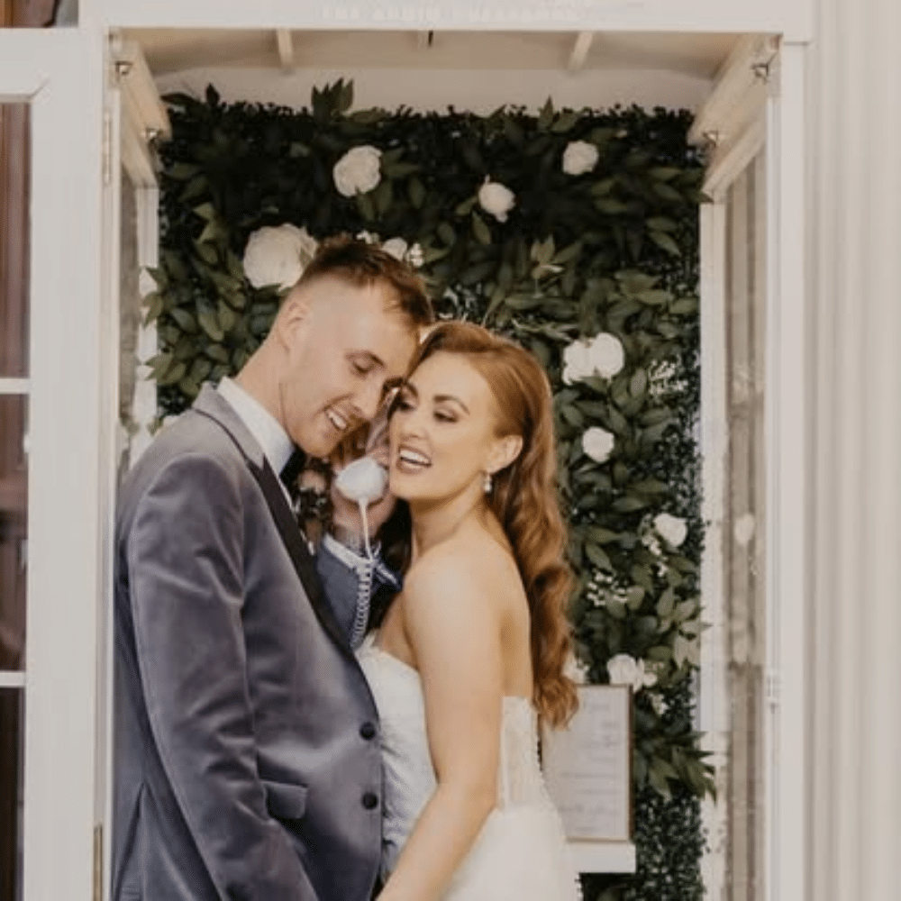 Bride and groom smiling in a floral-decorated photo booth.