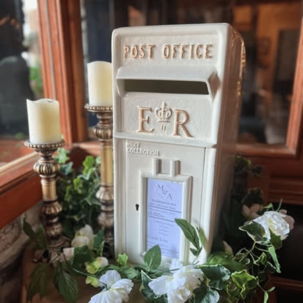 White postal box with "Post Office" and "E R" inscription, surrounded by candles and white flowers.