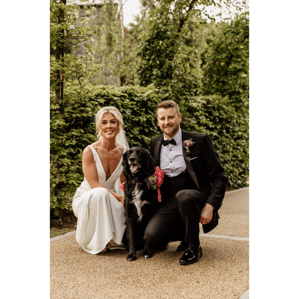 Bride and groom posing with a black dog, outdoors, on a path surrounded by greenery.