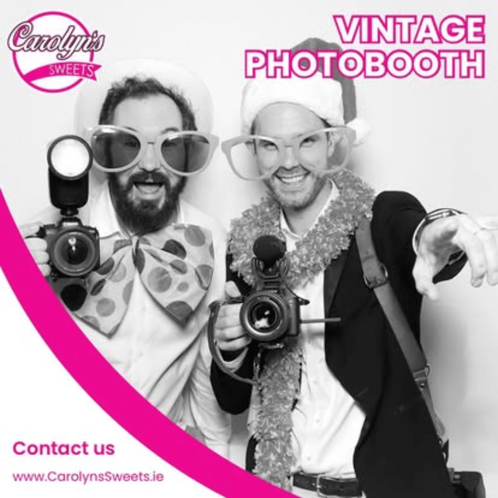 Two people in oversized glasses and festive attire posing with cameras in a vintage photobooth.