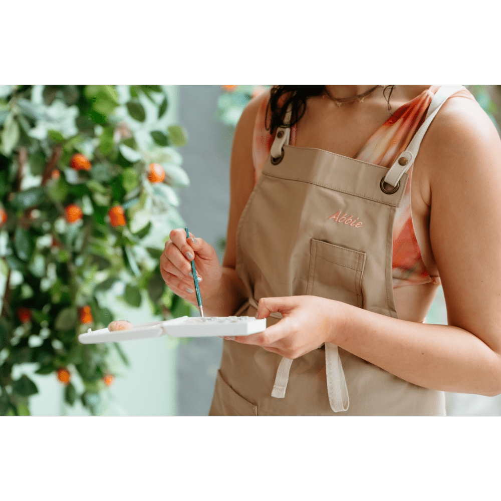 Person in apron holding a paint palette, painting, with green leafy background.