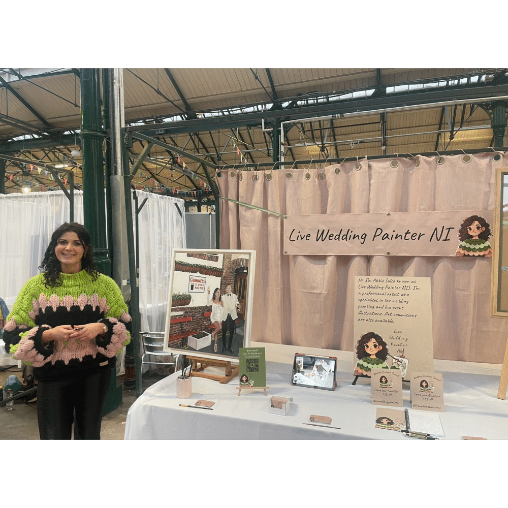 Live wedding painter exhibit with artist in colorful sweater, standing beside a display table, promotional materials, and sign.