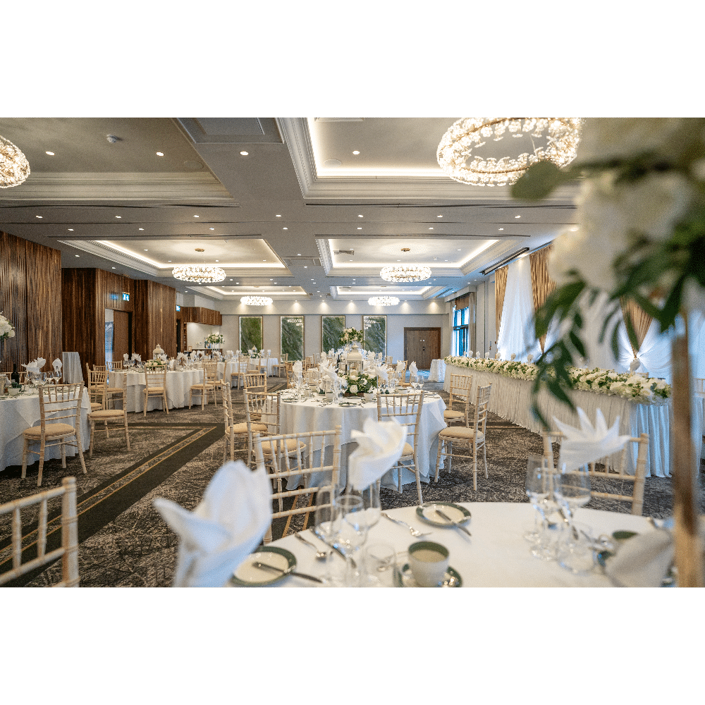 Elegant wedding reception setup with round tables, white linens, floral centerpieces, and chandeliers in a spacious venue.