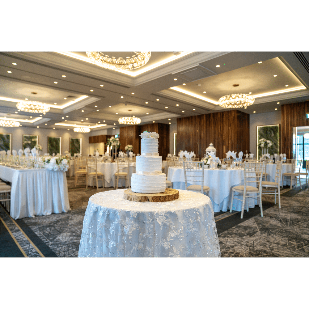 Elegant wedding reception setup with tiered cake on table, surrounded by decorated tables and chandeliers.