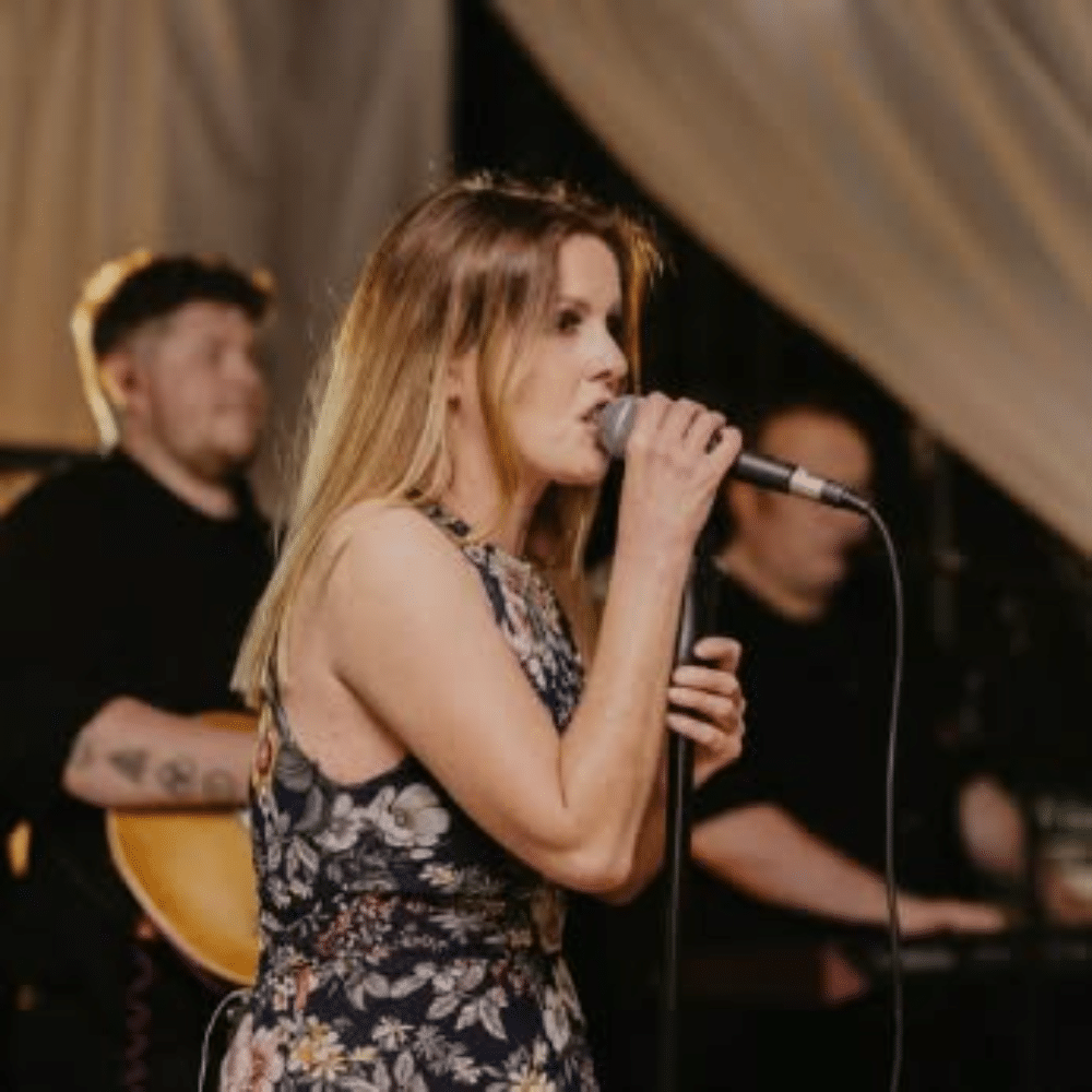 Woman singing with microphone on stage, guitarist and keyboardist in the background.