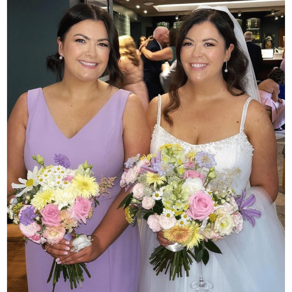 Bride and bridesmaid smiling with floral bouquets at a wedding.