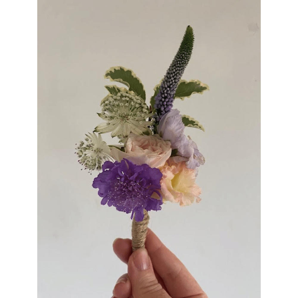 Hand holding a small bouquet with purple, pink, and white flowers on a light background.