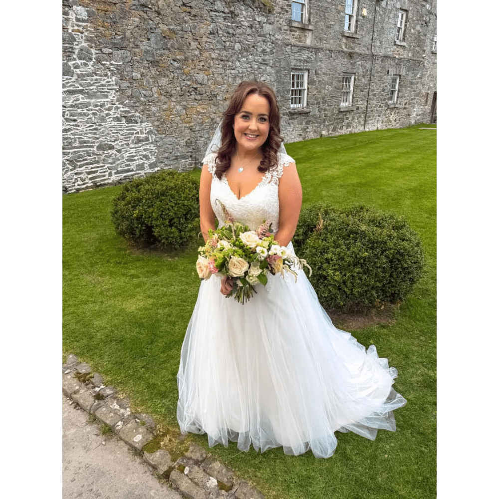 Bride in a white wedding dress holding a bouquet, standing on a manicured lawn by a stone building.
