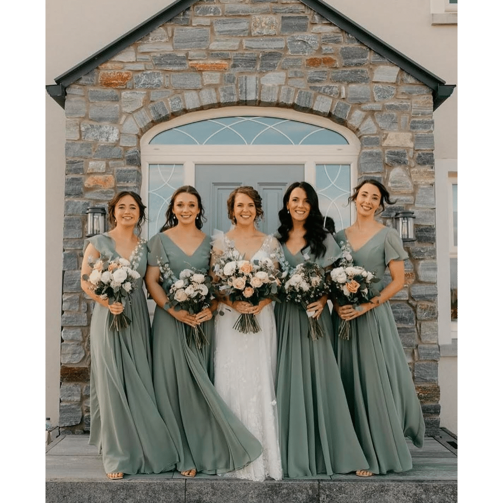 Bride and bridesmaids in green dresses holding bouquets, standing in front of a stone building with an arched door.