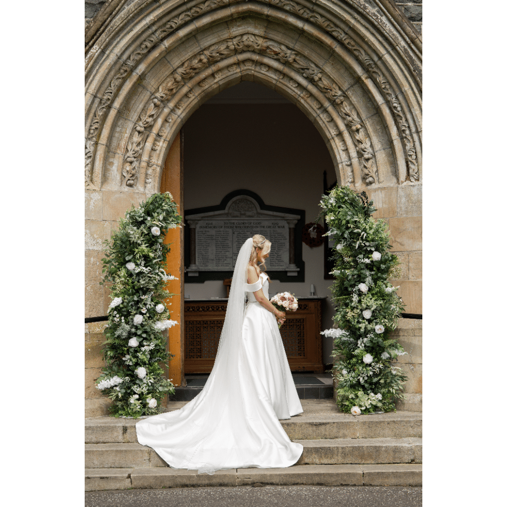Bride in white gown and veil holding bouquet, standing at ornate church entrance with floral decorations.