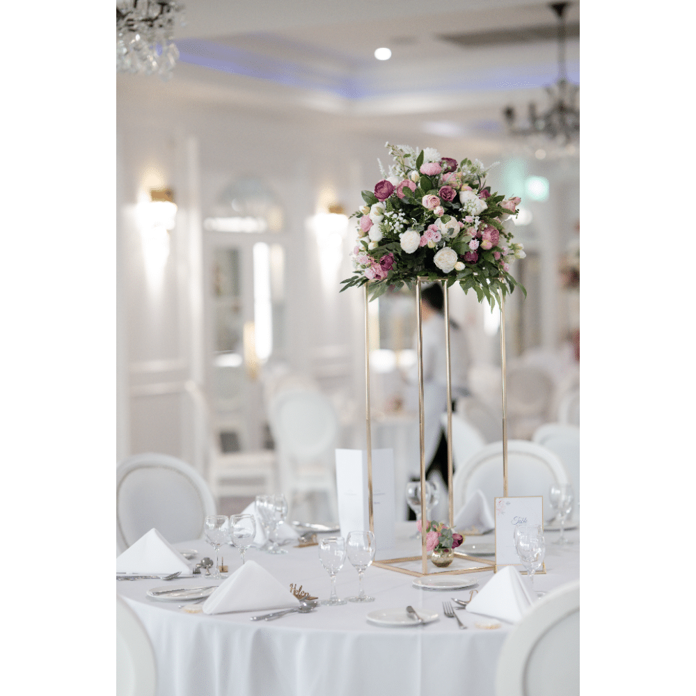 Elegant wedding table setting with tall floral centerpiece in a bright, opulent banquet hall.