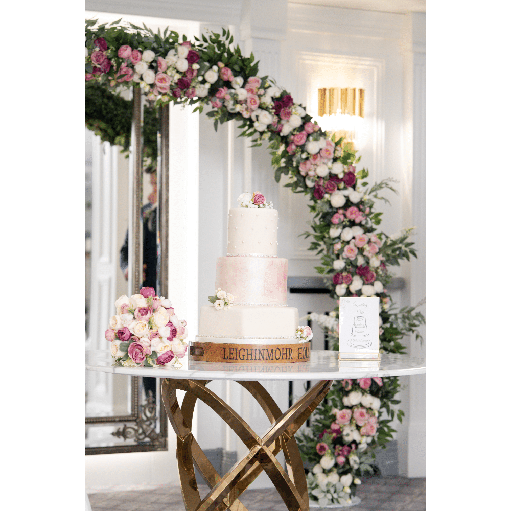 Elegant three-tier wedding cake with pink and white flowers, set on a decorative table with a floral arch in the background.
