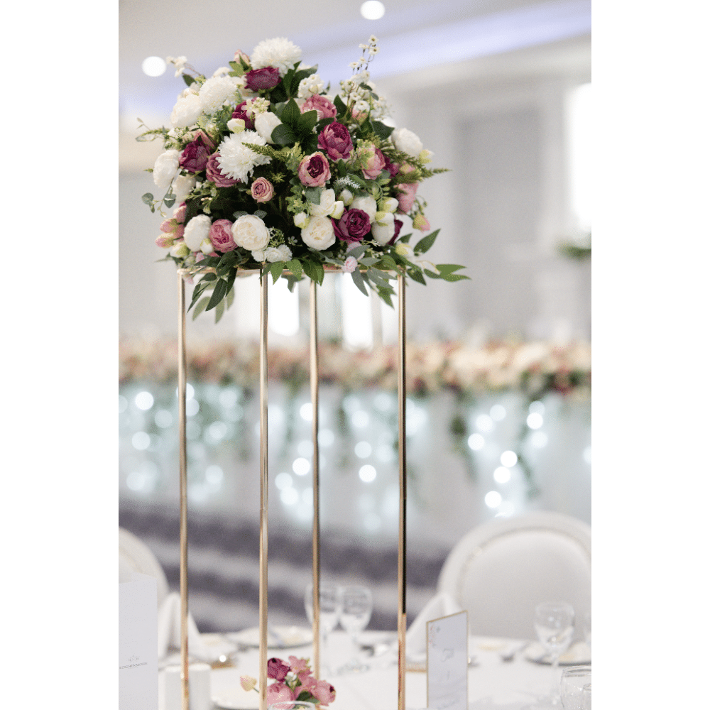 Elegant floral centerpiece on a tall stand with roses and greenery, set on a banquet table with blurred background.