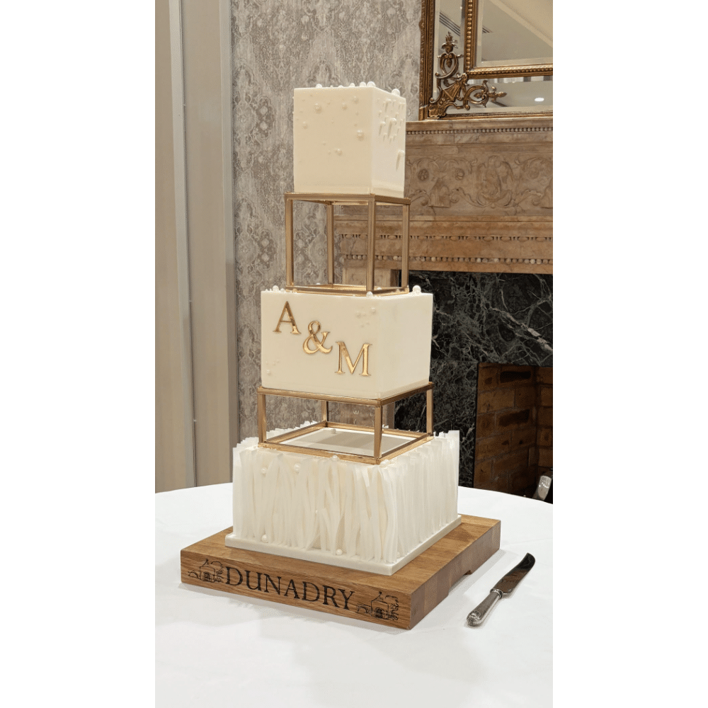 Tiered wedding cake with gold accents and initials "A & M," displayed on a wooden base labeled "Dunadry."