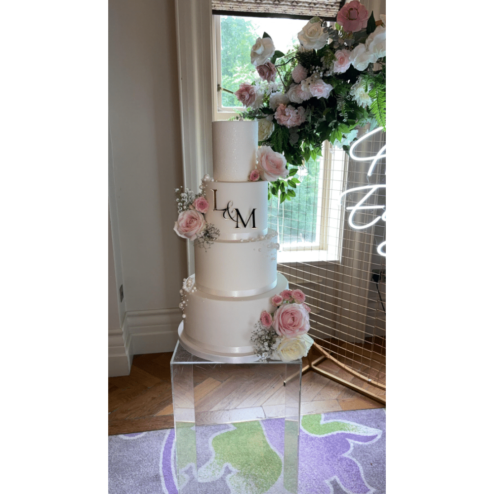 Elegant three-tier wedding cake with pink roses and "L&M" monogram on clear stand, beside floral arrangement.
