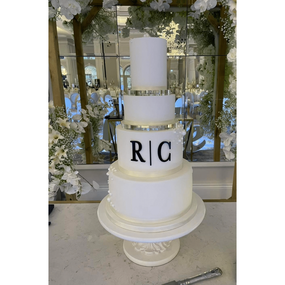 Elegant four-tier white wedding cake with initials "R | C" displayed on a decorative stand in a floral setting.
